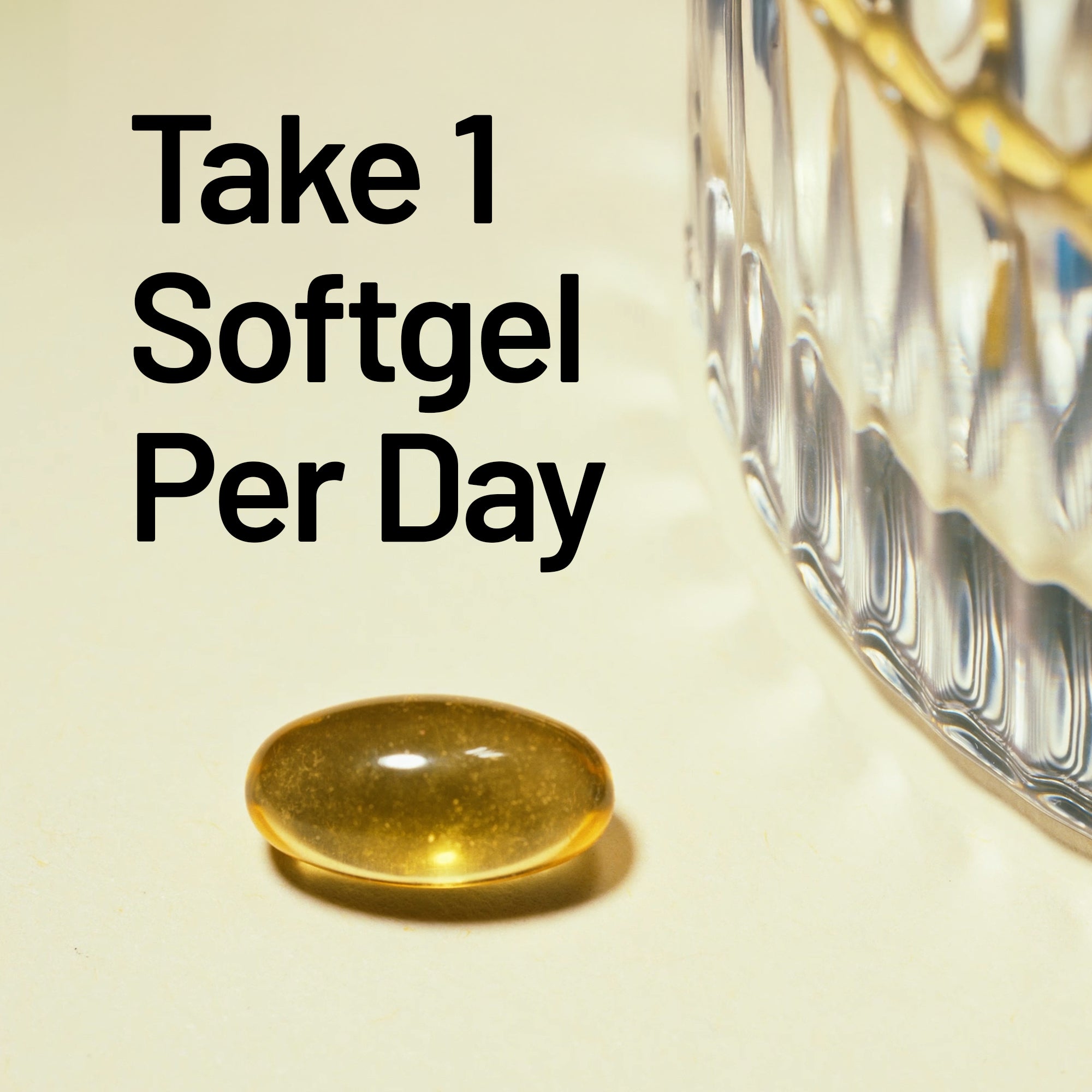 Gold softgel pill on a beige surface with text 'Take 1 Softgel Per Day' and a glass container in the background.