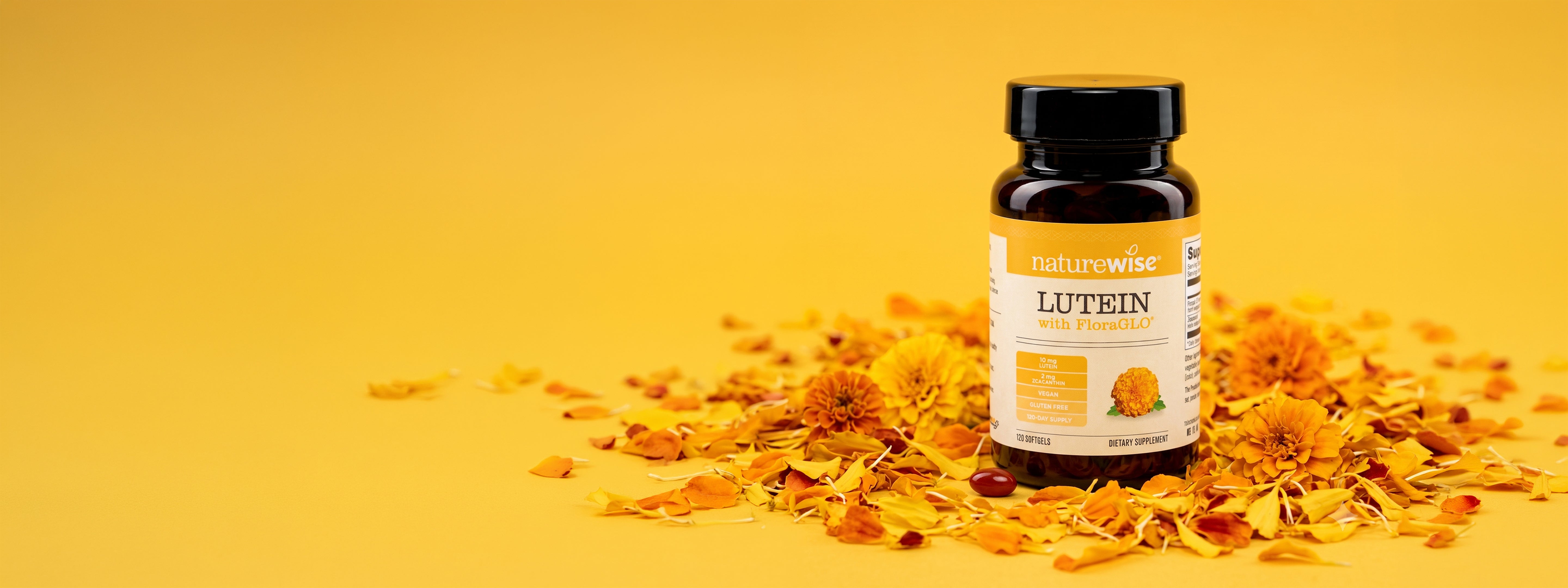 A brown bottle of NatureWise Lutein with FloraGLO dietary supplement stands upright on a bright yellow surface. The bottle is surrounded by scattered yellow and orange marigold petals and whole flower heads. A single dark red softgel capsule lies on the petals directly in front of the container.