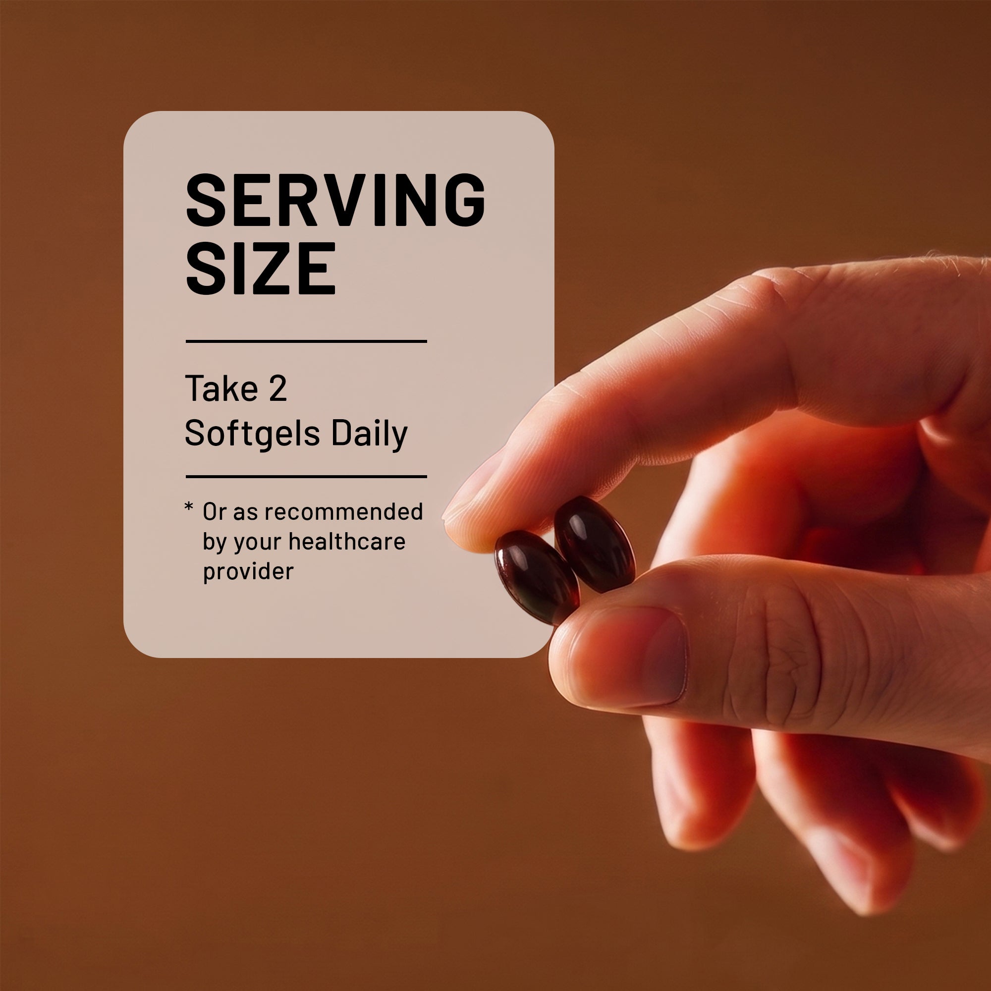 A hand holds two dark softgel capsules in front of a beige card that reads 'SERVING SIZE: Take 2 Softgels Daily' on a warm brown background.