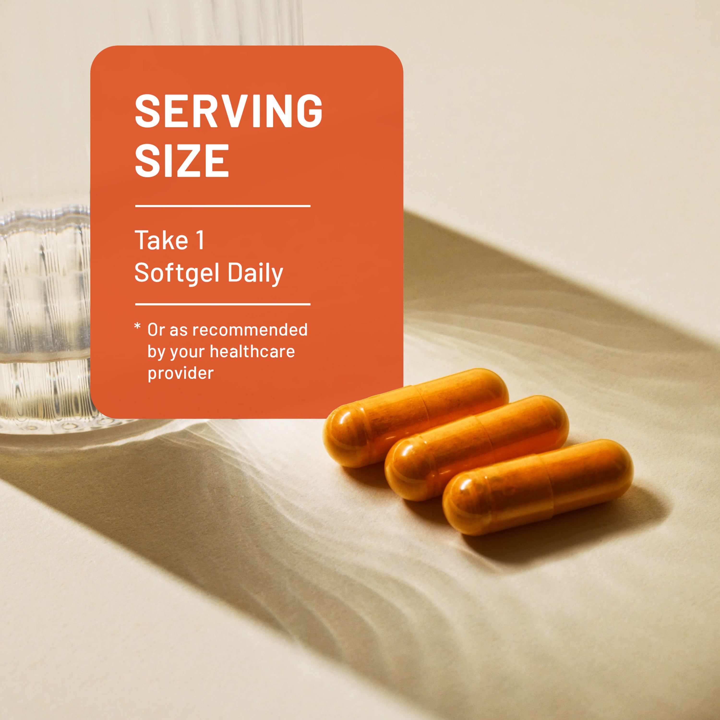 Three orange softgel capsules on a beige surface with a translucent glass, next to an orange label card that reads 'SERVING SIZE: Take 1 Softgel Daily'.