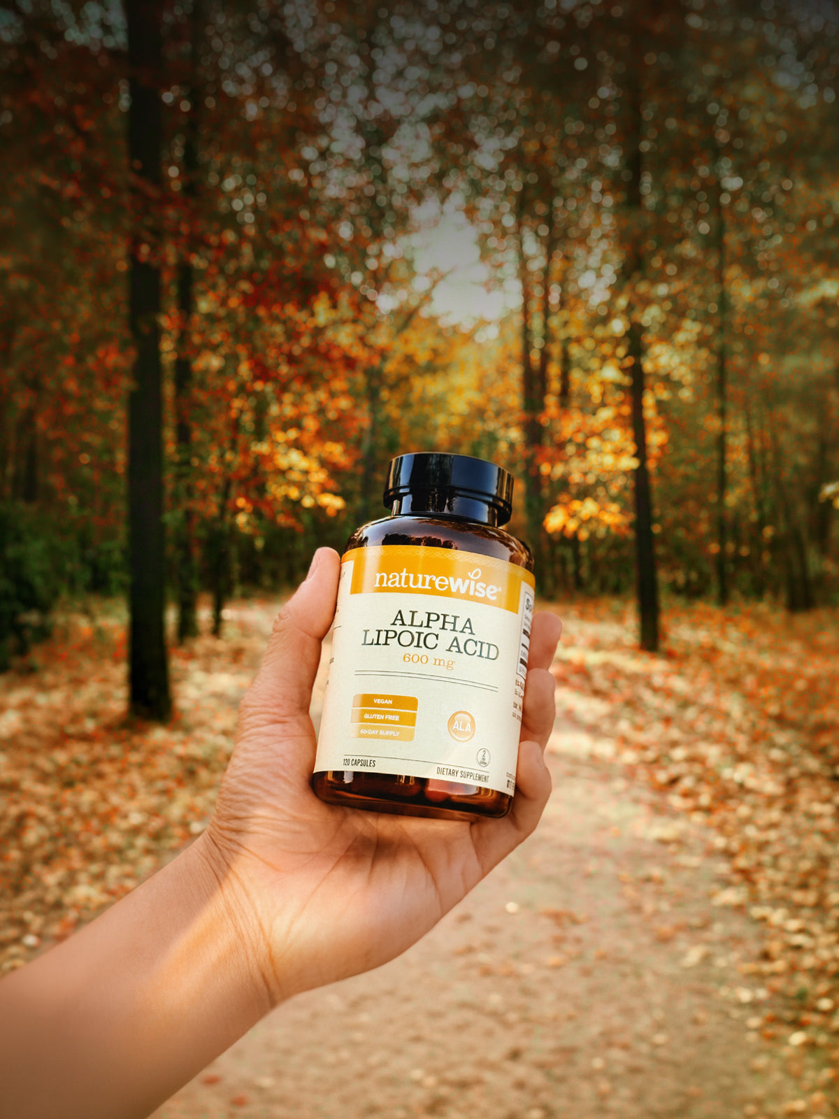 A hand holds a bottle of natureWise Alpha Lipoic Acid in an outdoor autumn forest setting with fallen leaves and trees in the background.