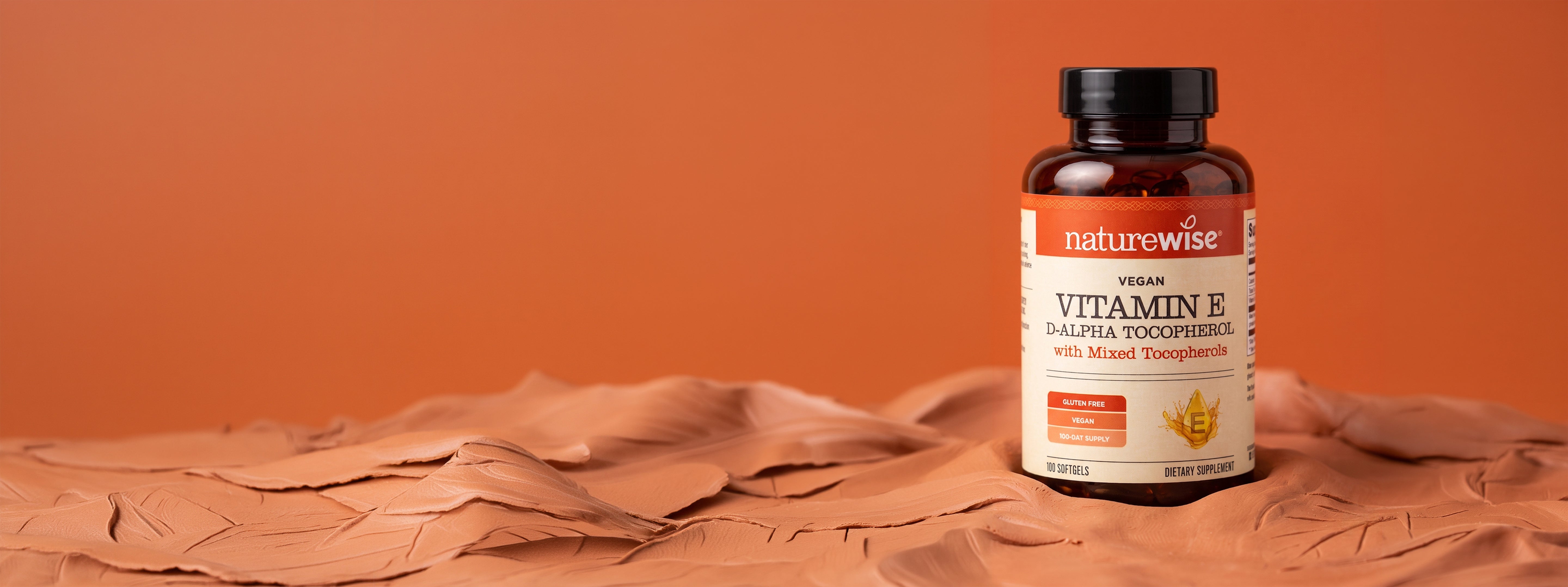 A bottle of Naturewise Vegan Vitamin E D-alpha Tocopherol with mixed tocopherols, placed on a sculpted clay surface against a solid terracotta background.