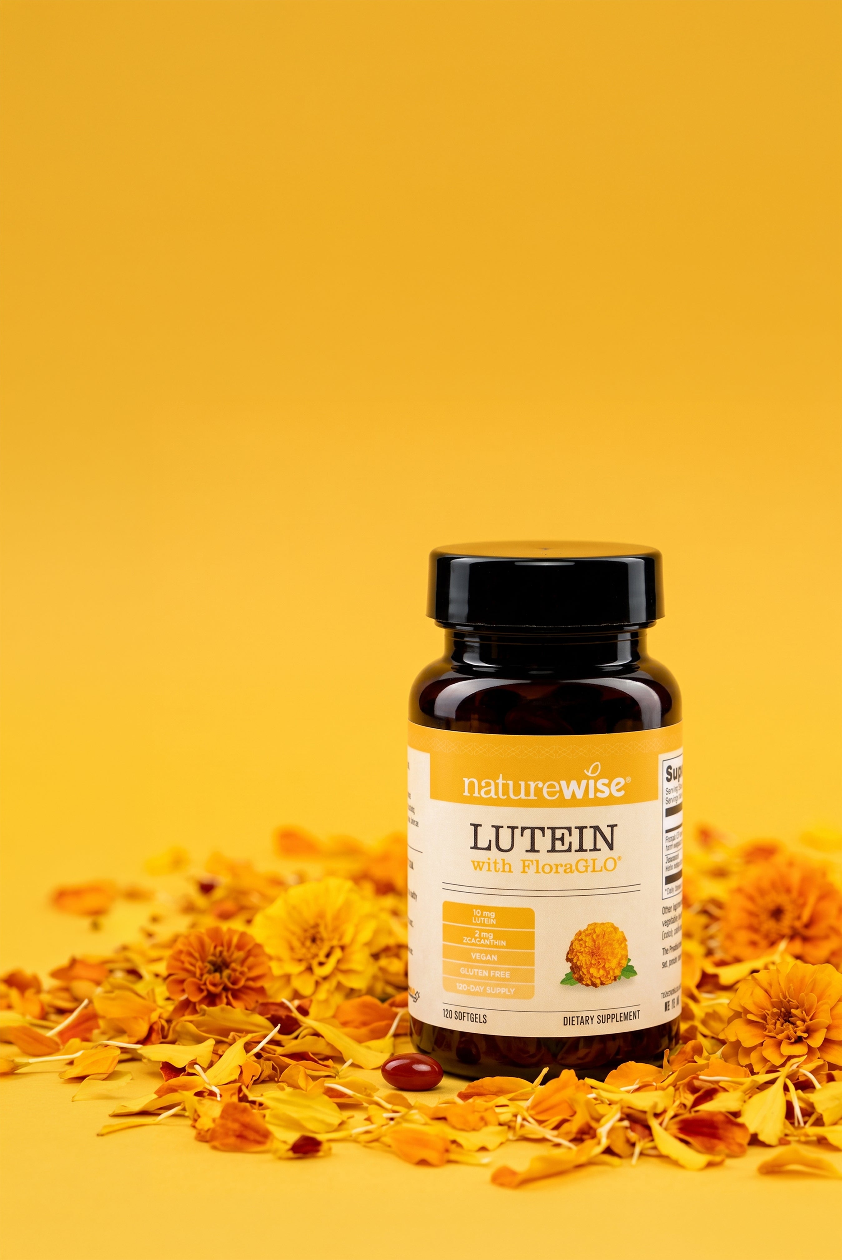 A brown bottle of NatureWise Lutein with FloraGLO dietary supplement stands upright on a bright yellow surface. The bottle is surrounded by scattered yellow and orange marigold petals and whole flower heads. A single dark red softgel capsule lies on the petals directly in front of the container.