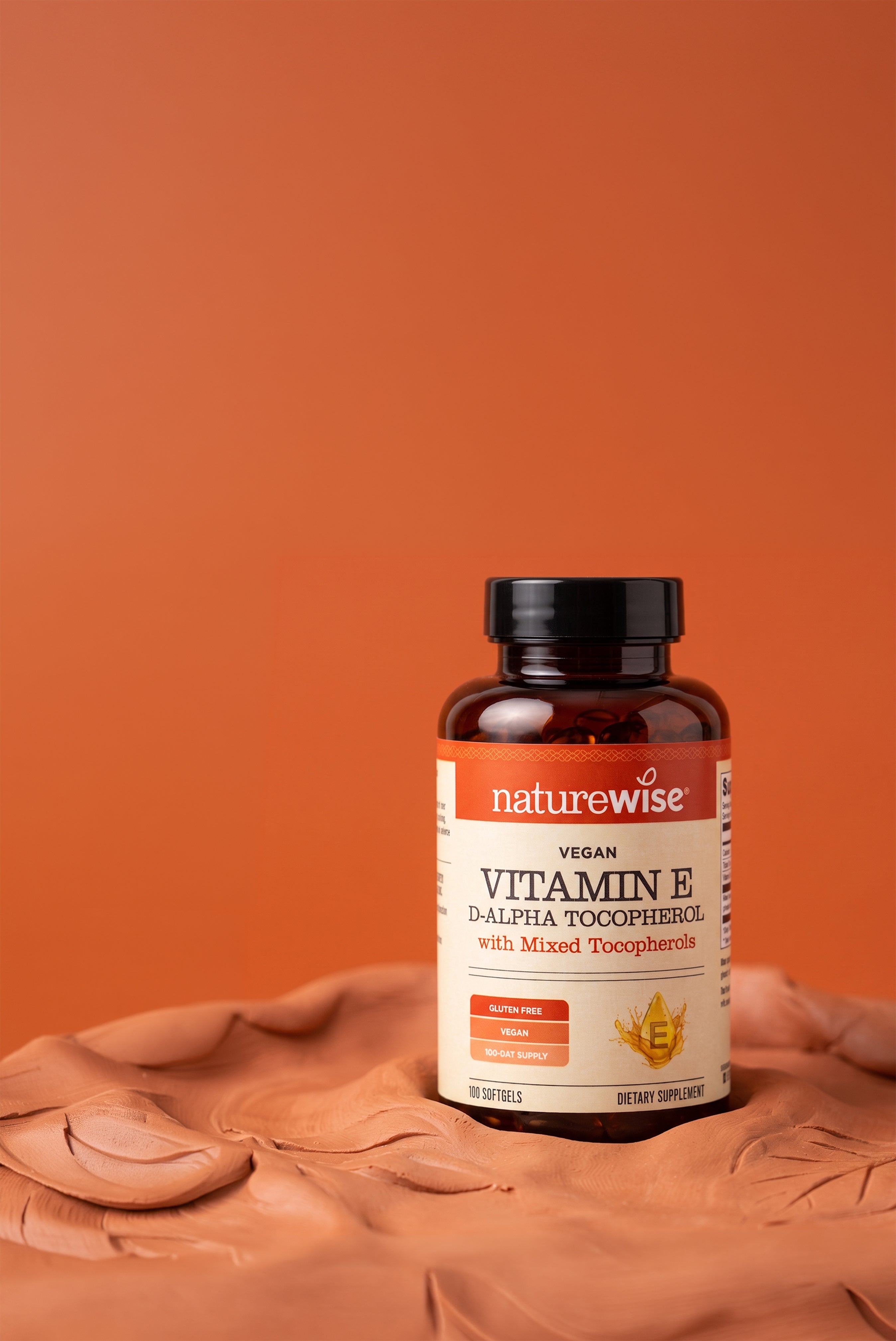 A bottle of Naturewise Vegan Vitamin E D-alpha Tocopherol with mixed tocopherols, placed on a sculpted clay surface against a solid terracotta background.